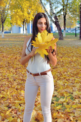 Beautiful girl in autumn park holding a maple leaf in her hand, smiling on her face, in white jeans and a blouse, against a background of trees and yellow foliage