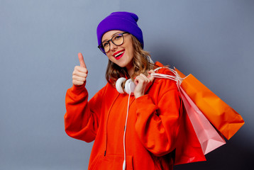 Style girl in orange hoodie and purple hat with shopping bags on grey background