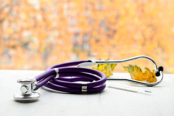 Stethoscope, thermometer and yellow leaves on white doctor's table, with autumn colors outside the...