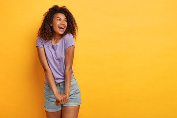 Joyful curly haired woman keeps hands together, laughs happily, focused aside, wears purple t shirt...