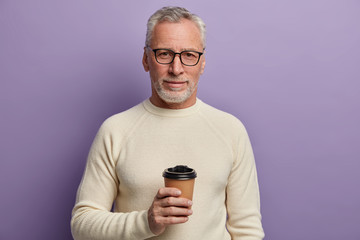 Grey haired senior man wears transparent glasses and white sweater, stands and cools hot beverage,...