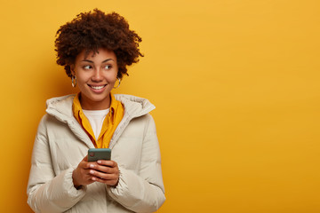 Image of thoughtful young Afro American woman watches live stream online, enjoys pleasant messaging in chat, poses against yellow background in winter outerwear, looks on right side with smile