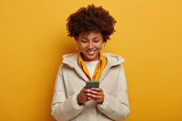Positive curly woman creats text publication via smartphone, being confident internet user, checks...