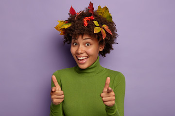 Horizontal shot of smiling lady with happy expression, points finger gun gesture into camera, wears green poloneck, has autumn leaves in hair, poses against purple background. You are best type