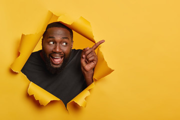 Photo of happy cheerful black male model points index finger aside, keeps mouth opened, shows tongue, attracts your attention to blank space, invites you going there, recommends product. Advertisement