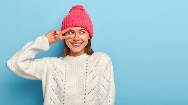 Upbeat glad woman looks playfully, makes peace gesture over eye, grins happily, enjoys amusing time, wears winter clothing, looks away, isolated over blue background, relaxes during weekend.
