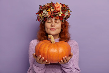 Horizontal shot of pretty redhead woman closes eyes with pleasure, holds ripe orange pumpkin,...