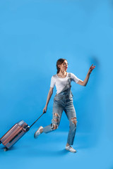 Fashionable emotional laughing and screaming girl running with suitcase on blue background. In a studio. Travel concept. Copy space.