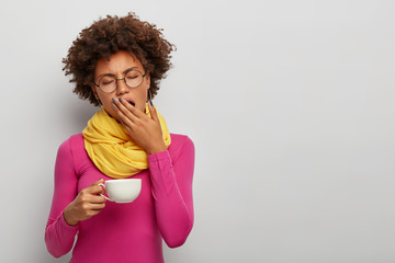 Tired curly woman yawn, has sleepy expression, drinks coffee early in morning, holds white muf of...