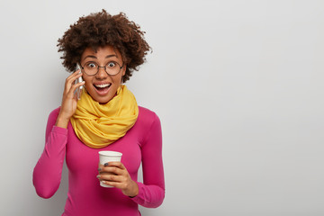 Joyous surprised African American woman gets awesome news during conversation via smartphone, has coffee break, looks with joy at camera through spectacles, wears bright clothing, poses indoor