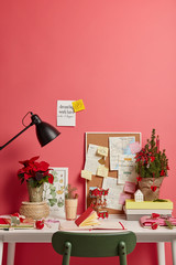 Holidays, interior, coworking space. Desktop with decorated fir tree, opened diary, wooden board on pink wall with written New Years resolutions on notes, written goals for Christmas. Eggnog to drink
