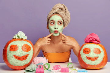 Pensive mixed race woman with organic clay mask, folds lips, has spa treatment, points both index finger at cheeks, wears wrapped towel on head, enjoys beauty procedures at home. Beautician client