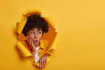 Shocked dark skinned female model looks aside with embarrassement, holds palm on cheekbone, surprised by big discounts, wears fashionable clothes, poses in paper hole, yellow studio background - obrazy, fototapety, plakaty