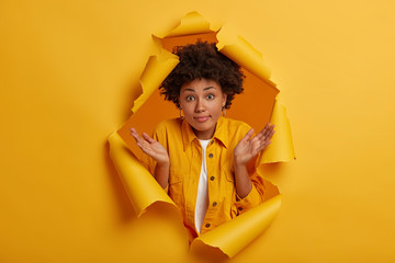 Uncertain cute woman with Afro hairstyle spreads palms, has confused clueless expression makes decision, shrugs shoulders has no idea poses in yellow paper hole background. How change life for better? - obrazy, fototapety, plakaty