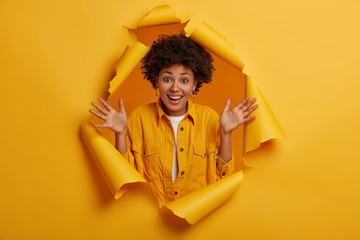 Indoor shot of joyful energetic woman with Afro hairstyle keeps palms raised, enjoys pleasant news, has natural curly hair, shows excitement and happiness, stands in ripped hole of yellow paper wall