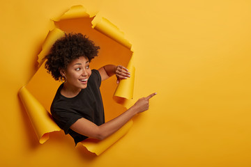 Joyous friendly looking smiling girl points aside with happy expression, toothy smile, pleased to...