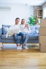 Young beautiful couple with dog sitting on the sofa drinking coffee at new home around cardboard boxes