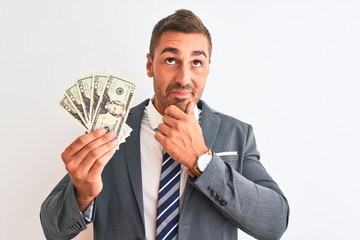 Young handsome business man holding bunch of dollars banknotes over isolated background serious...