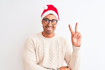 Young handsome man wearing christmas hat and glasses over isolated background smiling with happy face winking at the camera doing victory sign. Number two.