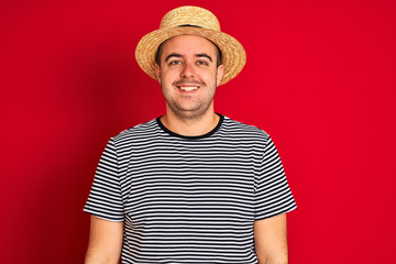 Young man wearing striped navy t-shirt and hat standing over isolated red background with a happy and cool smile on face. Lucky person.