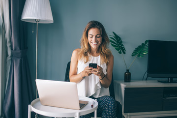 Happy female blogger in casual wear working remotely at home interior enjoying online chatting during break, cheerful Caucasian freelancer networking websites via modern smartphone gadget - obrazy, fototapety, plakaty