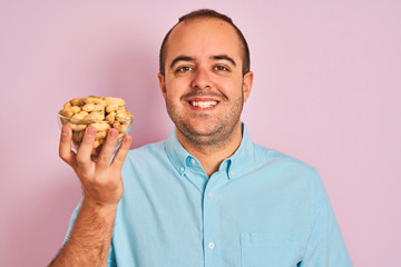 Young man holding bowl with peanuts standing over isolated pink background with a happy face standing and smiling with a confident smile showing teeth