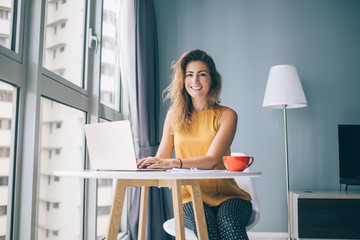 Portrait of happy housewife sitting at table in cozy apartment and spending leisure time for online shopping in web stores, positive female freelancer using home internet connection on laptop - obrazy, fototapety, plakaty
