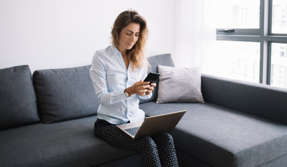 Young female chatting on phone sitting with laptop in apartment