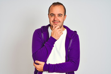 Young man wearing purple sweatshirt standing over isolated white background looking confident at the camera smiling with crossed arms and hand raised on chin. Thinking positive.