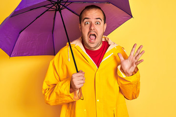 Young man wearing raincoat holding purple umbrella standing over isolated yellow background very...
