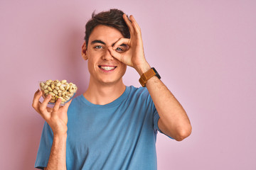 Teenager boy holding a bunch of pistachios over isolated pink background with happy face smiling doing ok sign with hand on eye looking through fingers