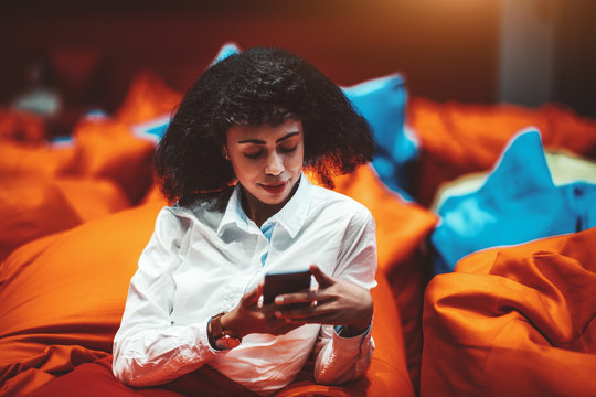 Beautiful African-American businesswoman is relaxing on an orange cushion in a lounge area of her office and using a smartphone; a biracial woman entrepreneur with a cellphone in a chillout zone