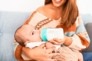 Young beautiful woman and her baby on the sofa at home. Newborn and mother relaxing and resting comfortable drinking milk using feeding bottle