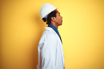 Afro american engineer man wearing white coat and helmet over isolated yellow background looking to side, relax profile pose with natural face with confident smile.