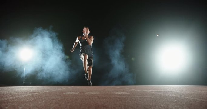 An asian male athlete is sprinting on stadium track during his evening training, spotted on smoked black background -sports concept 4k footage