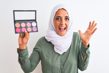 Young Arab woman wearing hijab showing makeup blush palette over isolated background very happy and excited, winner expression celebrating victory screaming with big smile and raised hands