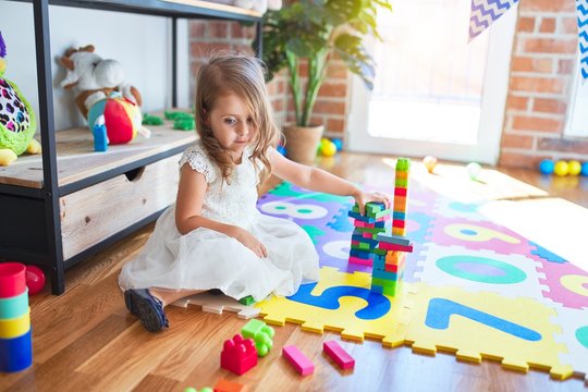 Adorable blonde toddler playing with building blocks toy around lots of toys at kindergarten