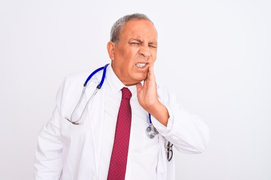 Senior grey-haired doctor man wearing stethoscope standing over isolated white background touching mouth with hand with painful expression because of toothache or dental illness on teeth