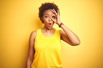 Beauitul african american woman wearing summer t-shirt over isolated yellow background doing ok gesture shocked with surprised face, eye looking through fingers. Unbelieving expression.