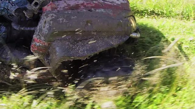 Dirty extreme quadrocycle is actively riding and moving along offroad with mud, grasses and bushes outside on sunny warm day.