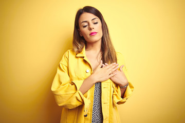 Young beautiful woman wearing denim jacket standing over yellow isolated background smiling with hands on chest with closed eyes and grateful gesture on face. Health concept.