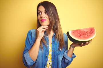 Young beautiful woman eating fresh healthy watermelon slice over yellow background serious face...