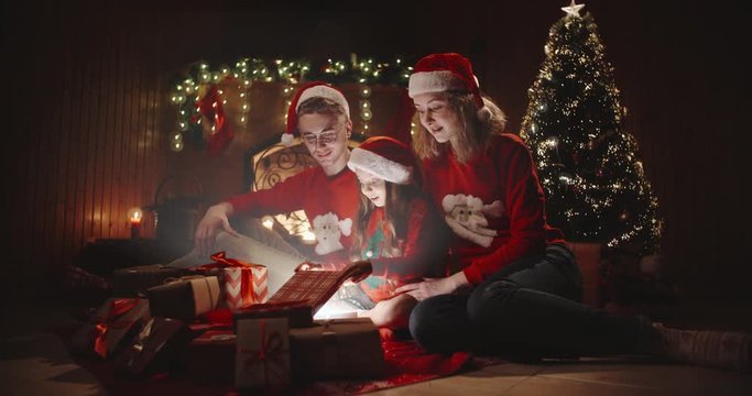 Caucasian family of three sitting near chimney, little girl opening up her magical gift that shines and sparkles - togetherness, love, christmas spirit concept 4k footage