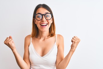 Beautiful redhead woman wearing glasses over isolated background celebrating surprised and amazed for success with arms raised and open eyes. Winner concept.