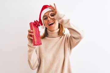 Beautiful redhead woman wearing christmas hat holding present over isolated background with happy face smiling doing ok sign with hand on eye looking through fingers