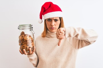 Beautiful redhead woman wearing christmas hat holding cookies jar over isolated background with...