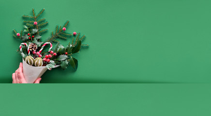 Christmas panoramic background flat lay on green paper with copy-space. Female hand holding veneer cone with fir and holly twigs with candy canes, dry fruit and berries.
