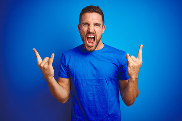 Young handsome man wearing casual t-shirt over blue isolated background shouting with crazy expression doing rock symbol with hands up. Music star. Heavy concept.