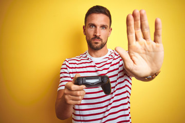 Young handsome man playing video games using joystick game pad over yellow background with open hand doing stop sign with serious and confident expression, defense gesture © Krakenimages.com