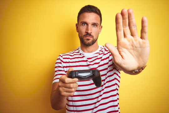Young handsome man playing video games using joystick game pad over yellow background with open hand doing stop sign with serious and confident expression, defense gesture